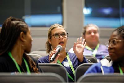 Student with microphone in seated room with hand up asking a question.