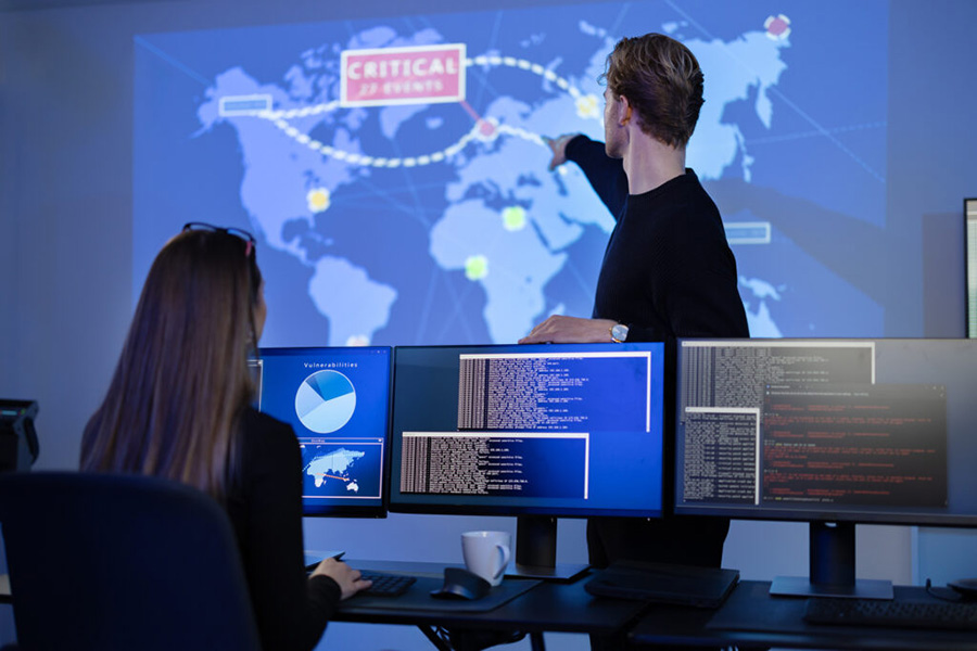 Two people in a computer room with one pointing at a projection on the wall with map of the world.