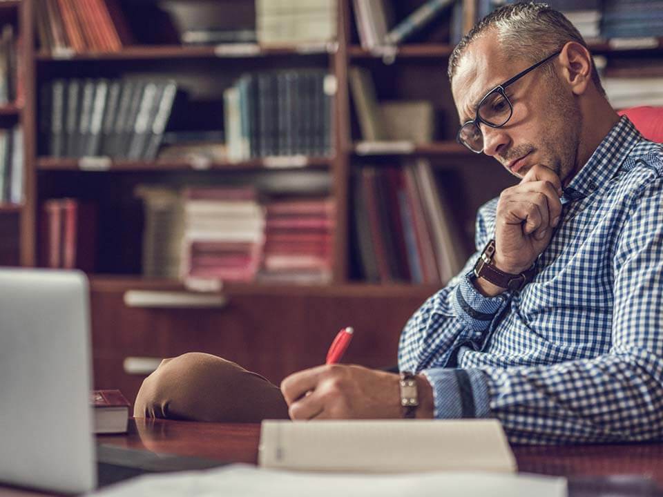 A man writing something on a notebook with an open laptop in front of him