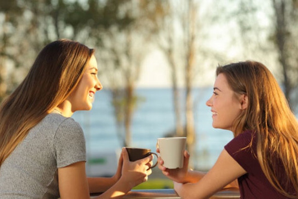 Two young women seated outdoor holding a cup and smiling at each other