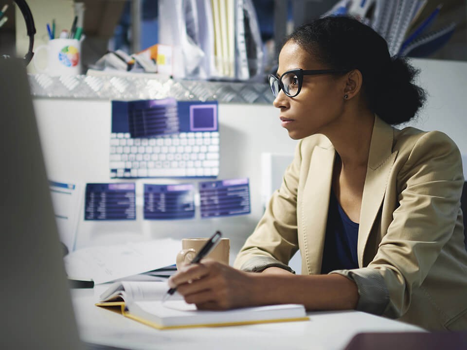 A woman with a pen in her hands and holding it above a notebook looking at something in front of her